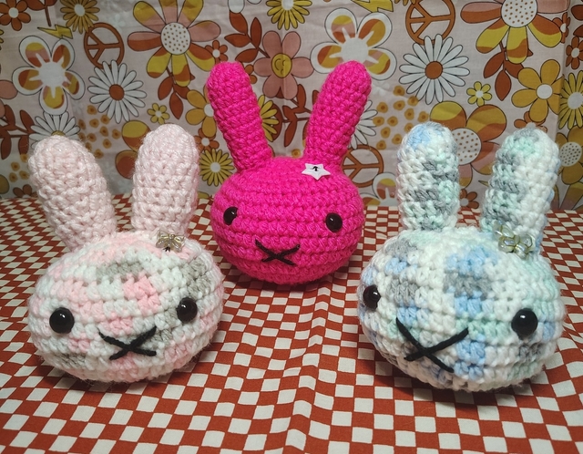 Three crocheted bunny plushies in pink, hot pink, and blue against a retro floral backdrop — handmade by Cosmic Pocket